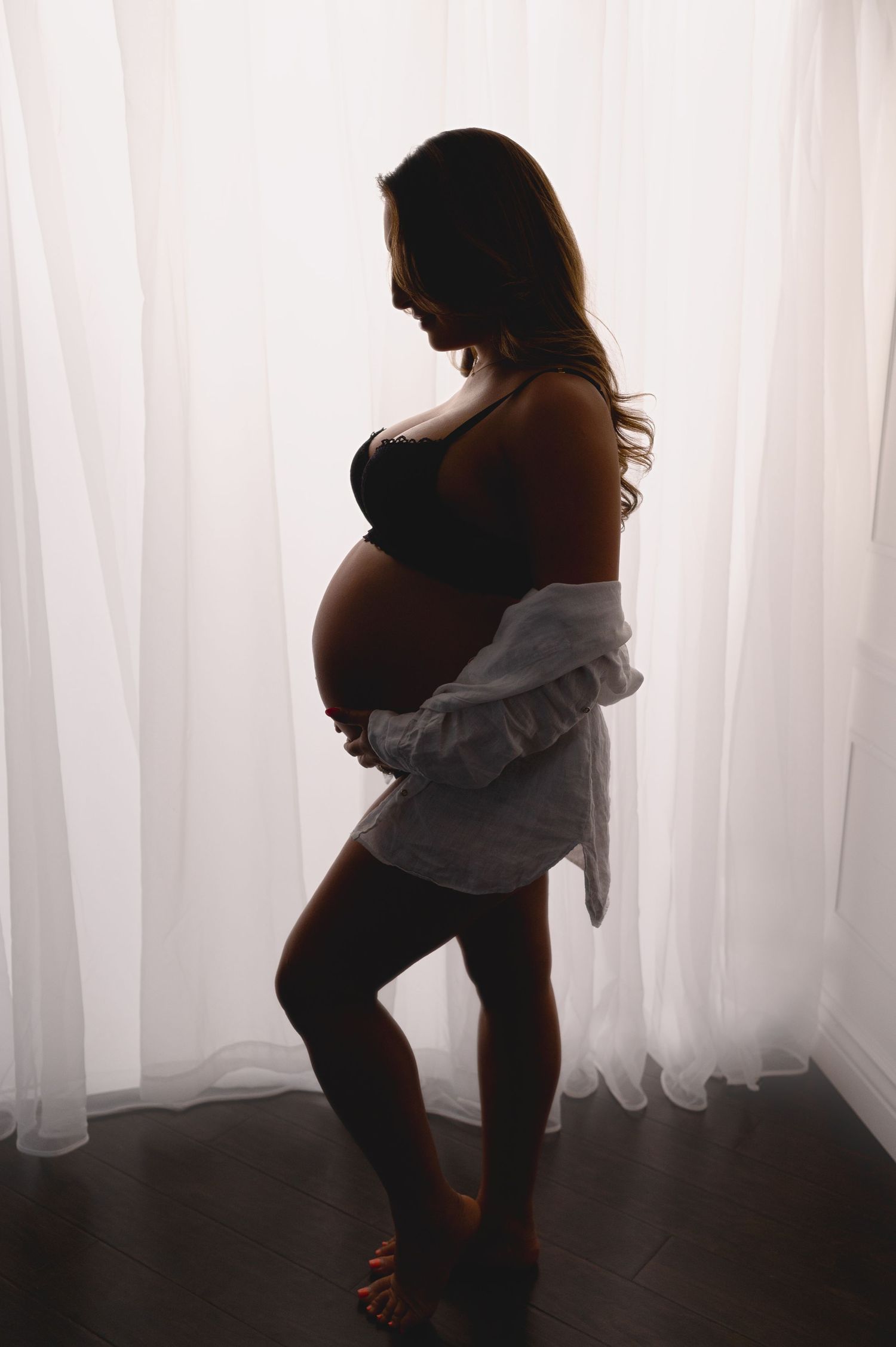 Silhouetted side view of pregnant woman in white shirt standing by window.