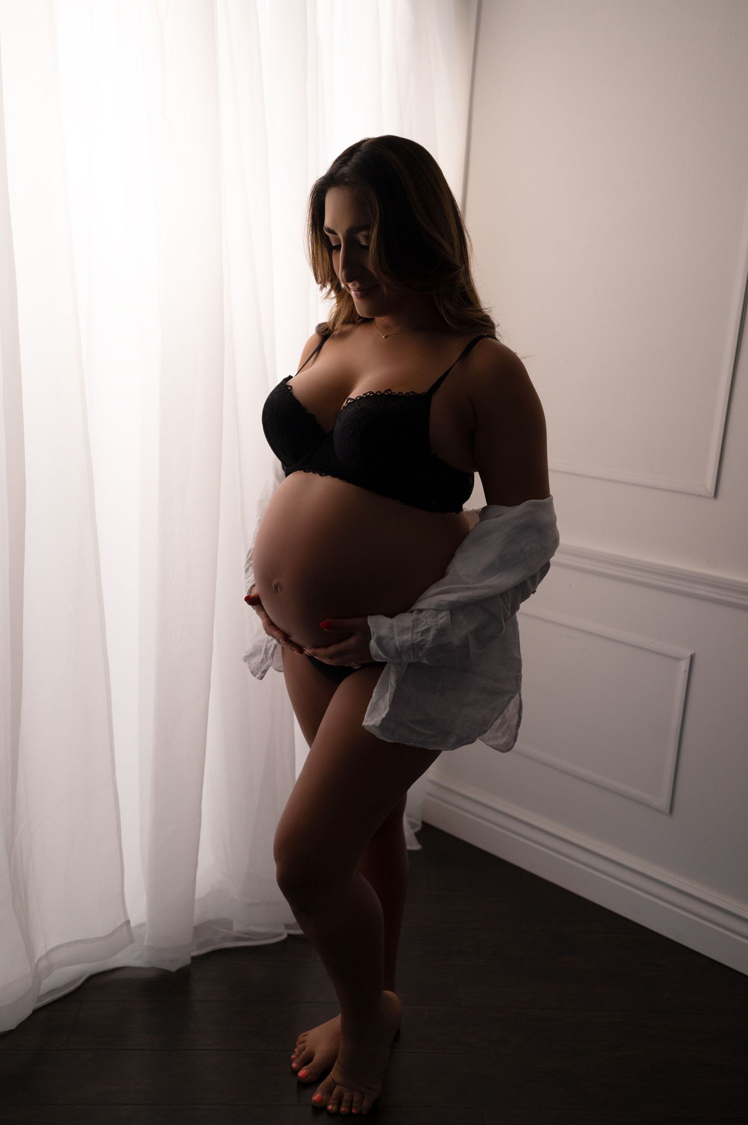 A maternity photo of a woman in black lingerie and open grey shirt against a white wall.