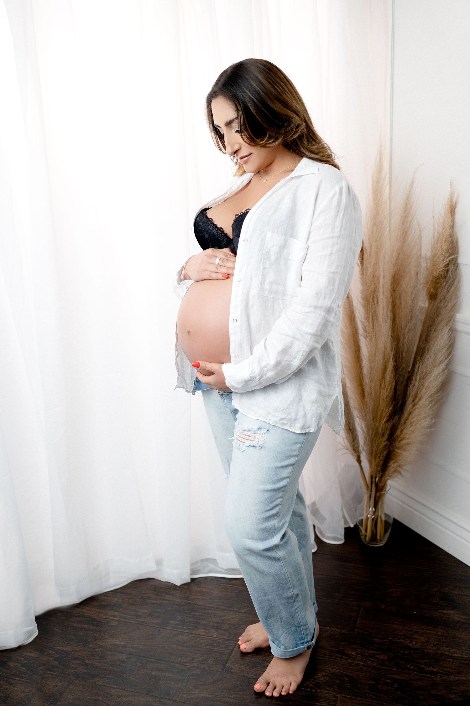 Full-length maternity photo of woman in white shirt and distressed jeans beside pampas grass decor.