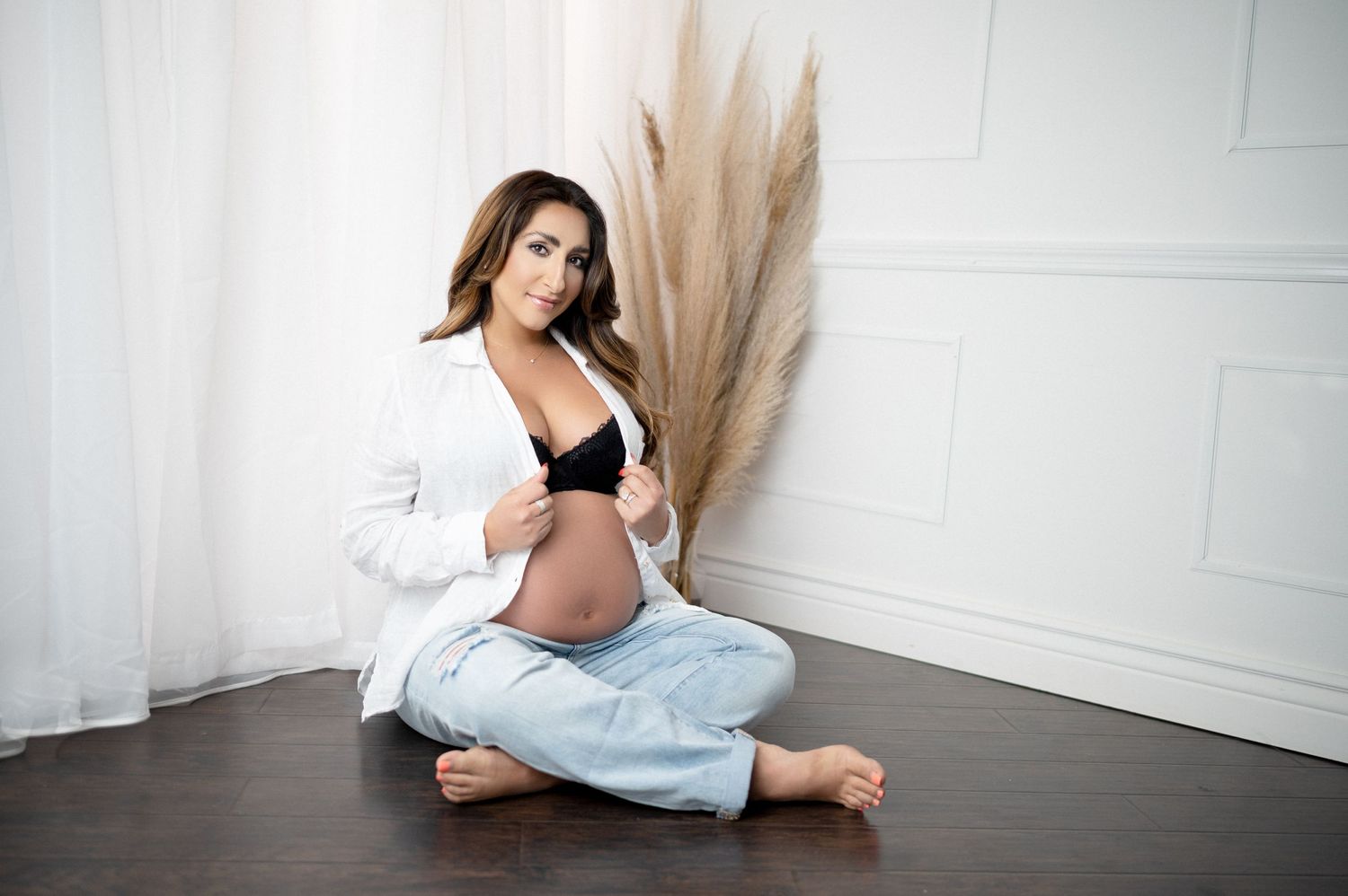 Woman in casual white shirt and jeans poses for maternity photo with pampas grass decor.