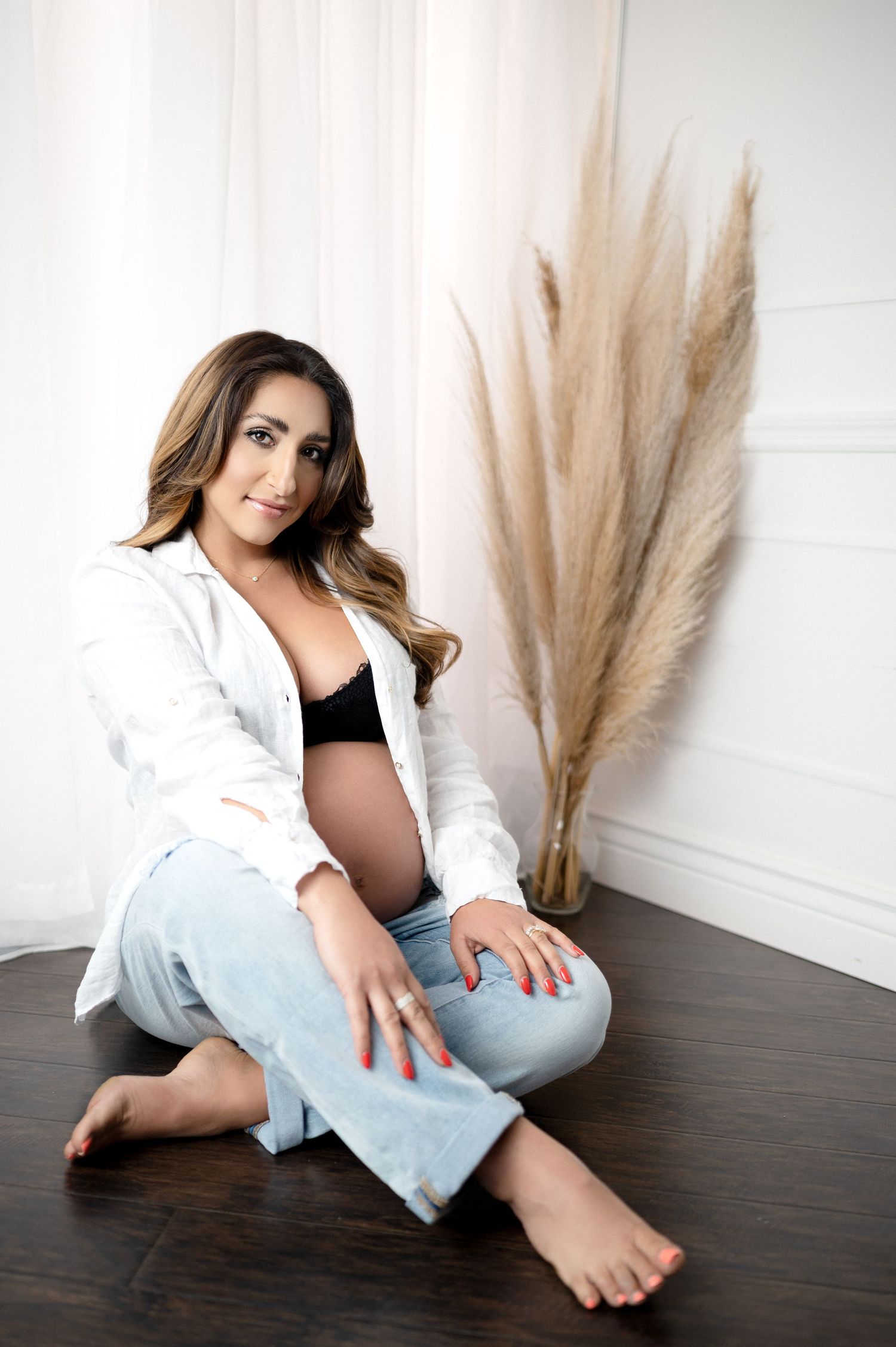 Expectant mother in white shirt and jeans sits on floor next to pampas grass in studio setting.