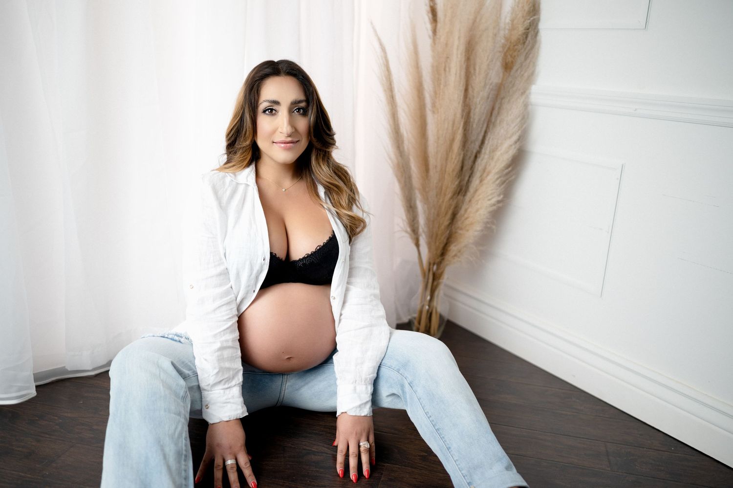 Maternity portrait with woman in white shirt and jeans sitting near decorative pampas grass.