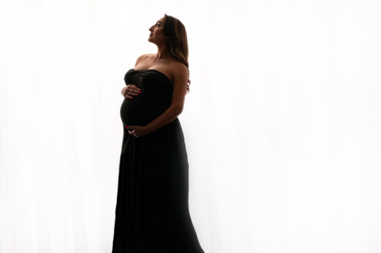 Silhouette maternity photo of woman in black dress against bright white background.