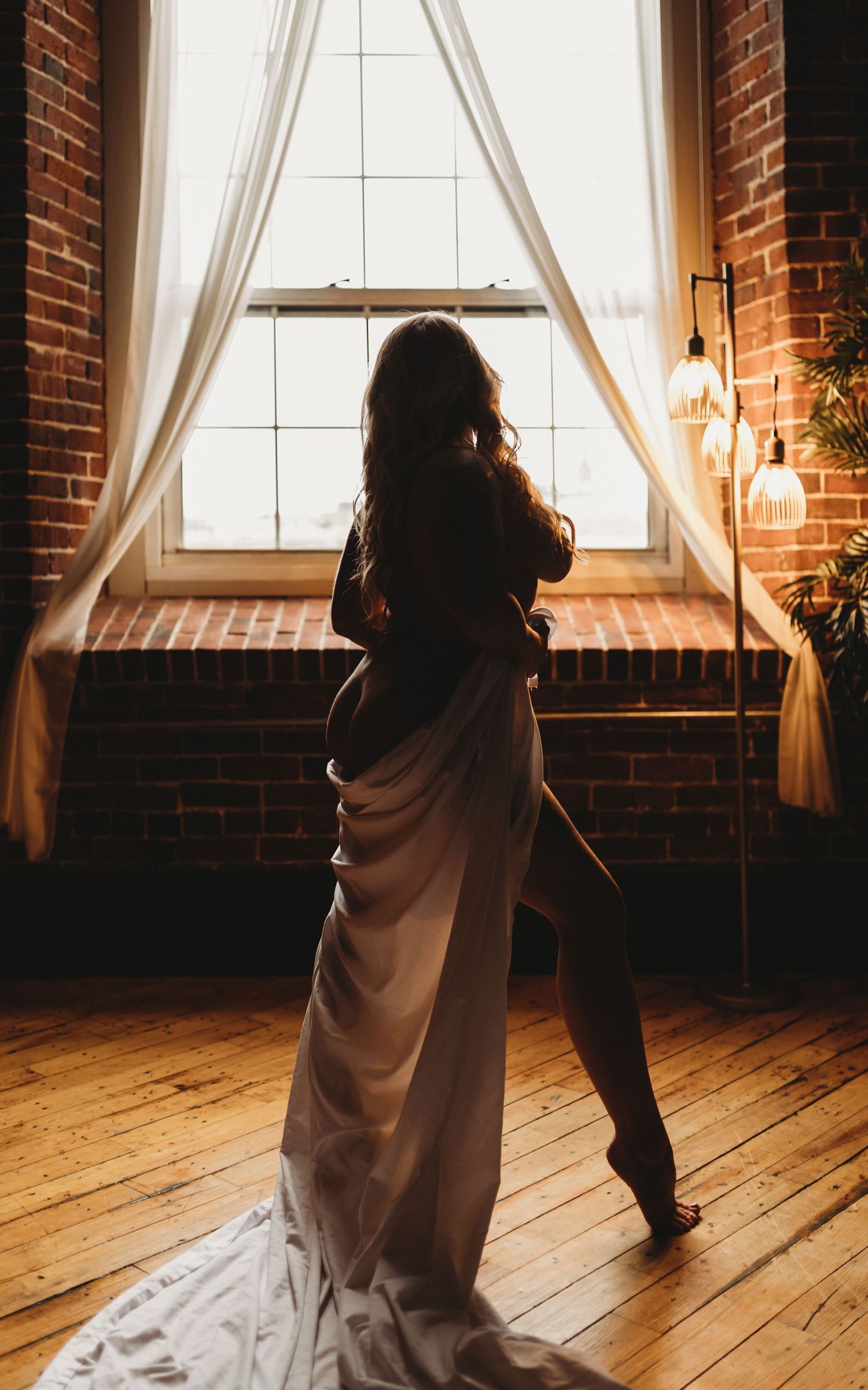 Silhouette figure by large window with flowing fabric and exposed brick walls.
