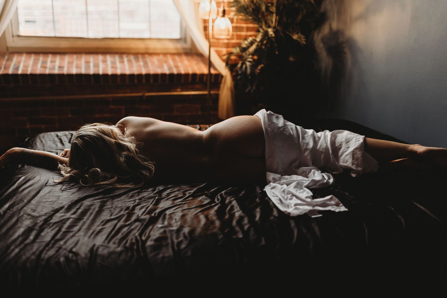 Sunlit bedroom with person sleeping peacefully on rumpled dark bedding in morning light.