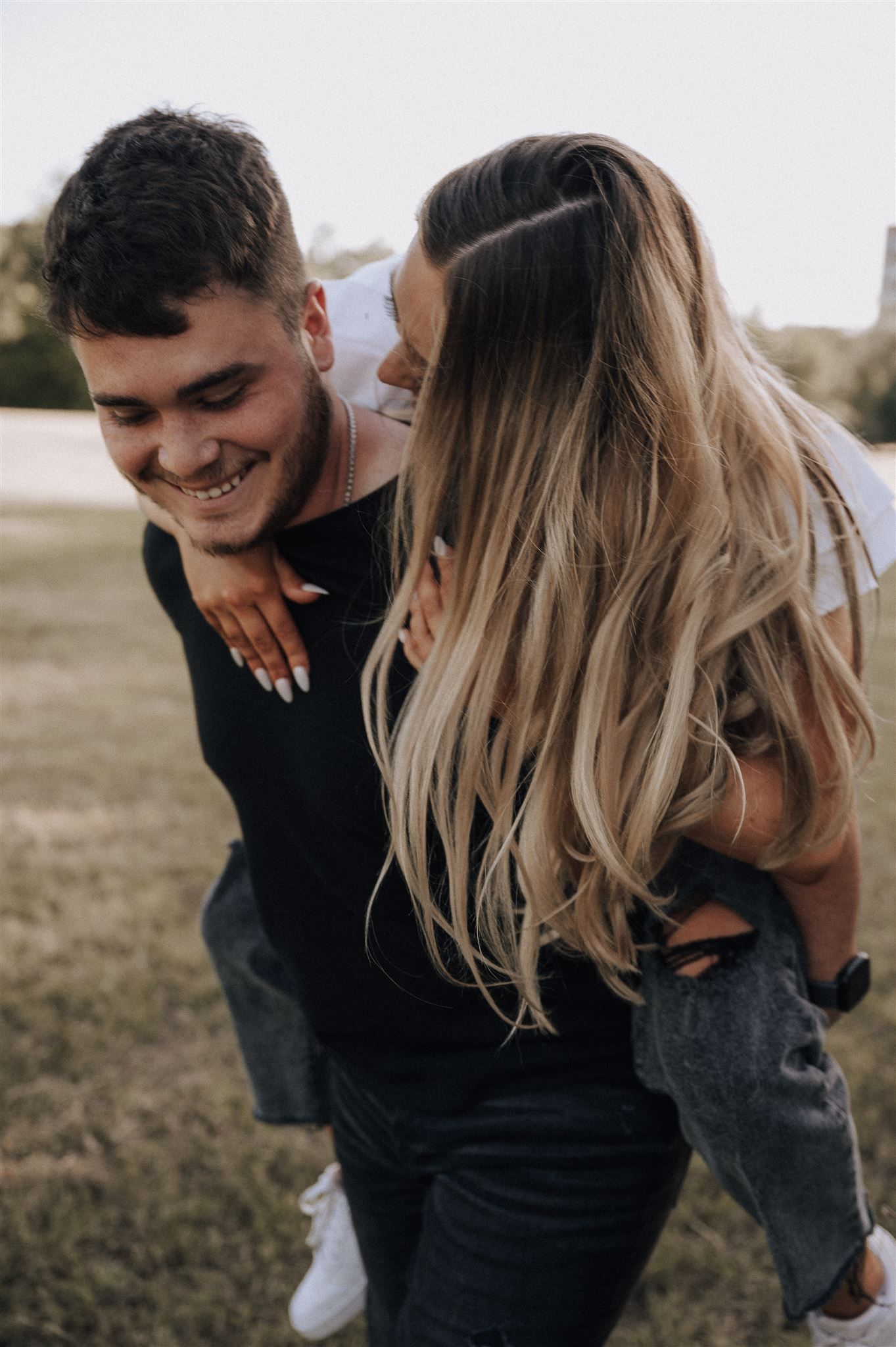 A couple shares a playful piggyback ride moment outdoors with a warm sunset glow.
