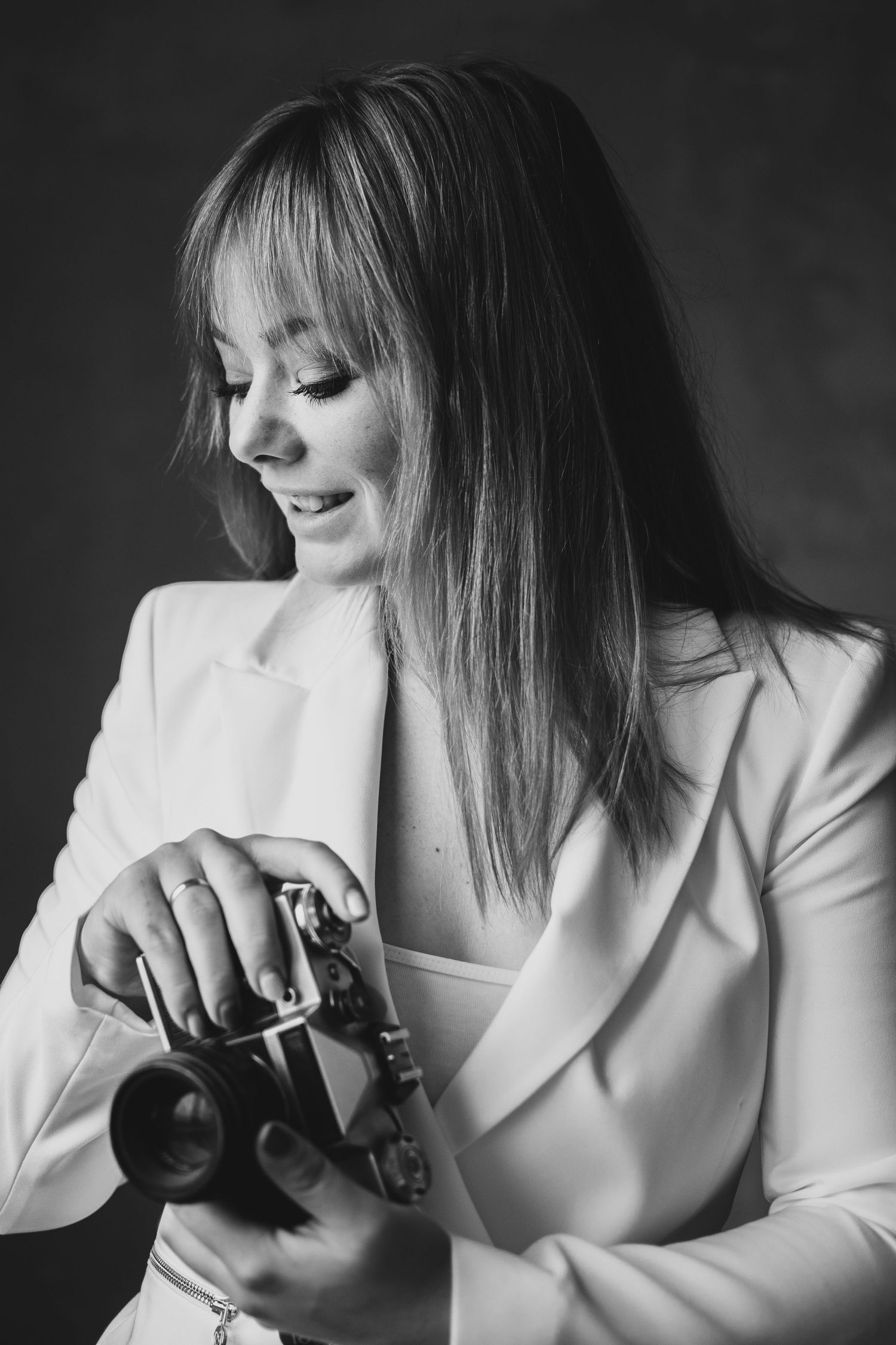 Artistic black and white portrait of someone in white blazer examining a camera with gentle expression.