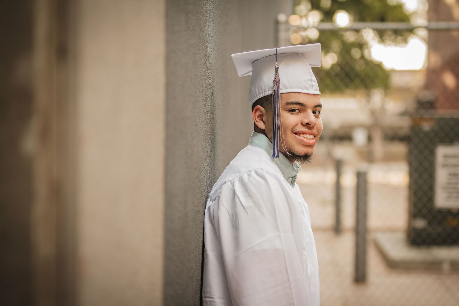 Graduate wearing white cap and gown poses outdoors with a bright smile.