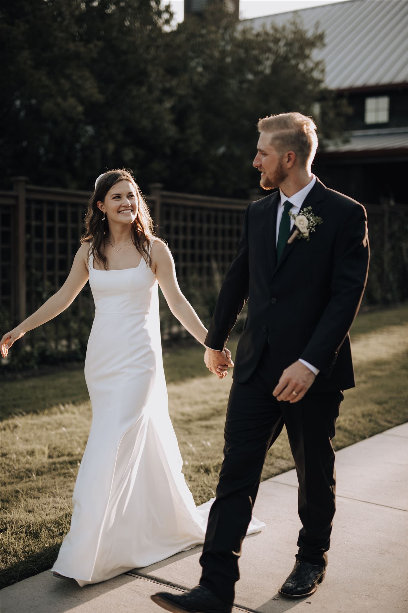 Happy couple walks holding hands along pathway with rustic building in background.