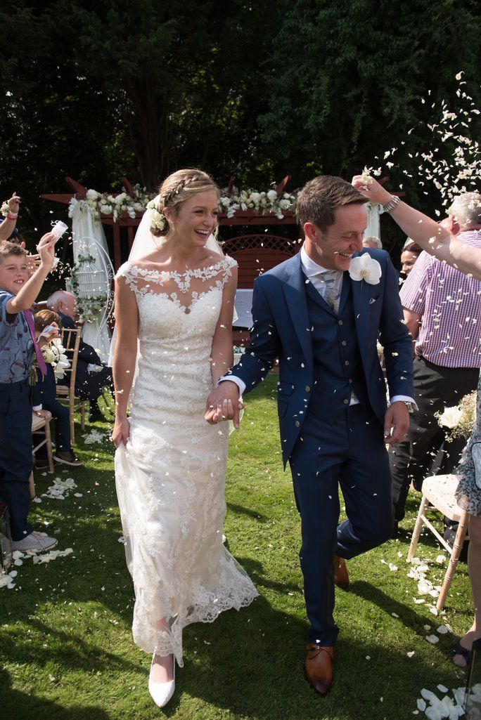 Newlyweds walk through shower of confetti while guests celebrate on sunny garden lawn.