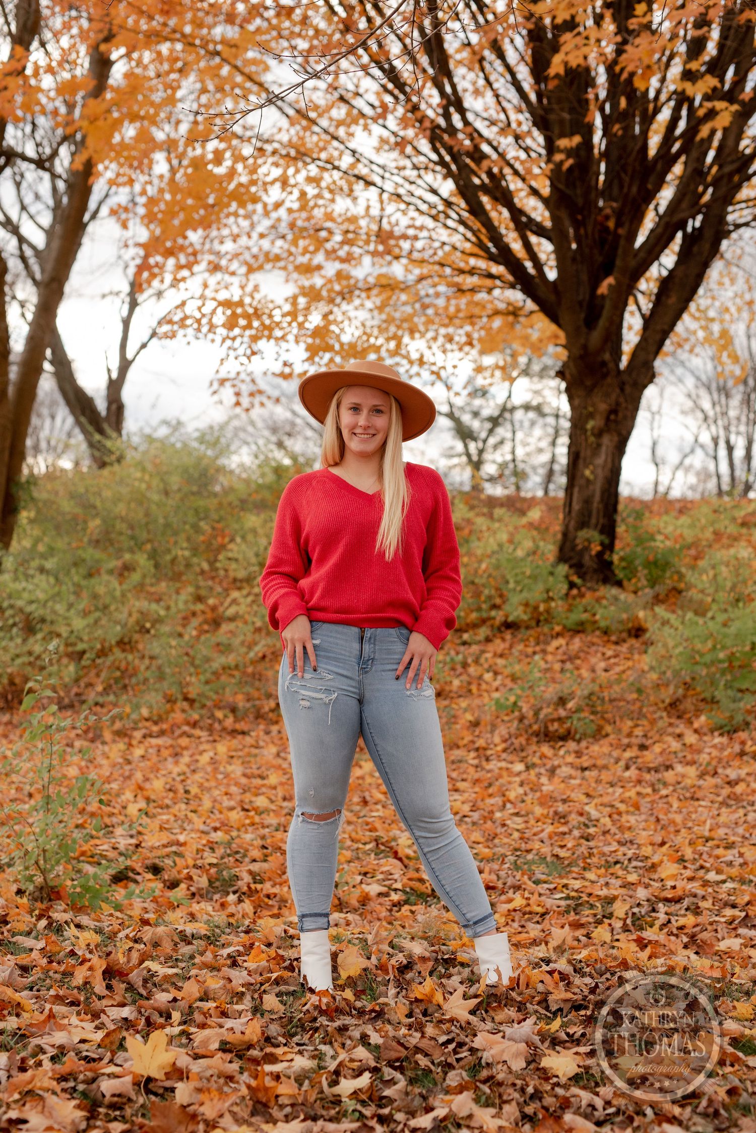 Someone in jeans and red sweater stands confidently among fallen autumn leaves.