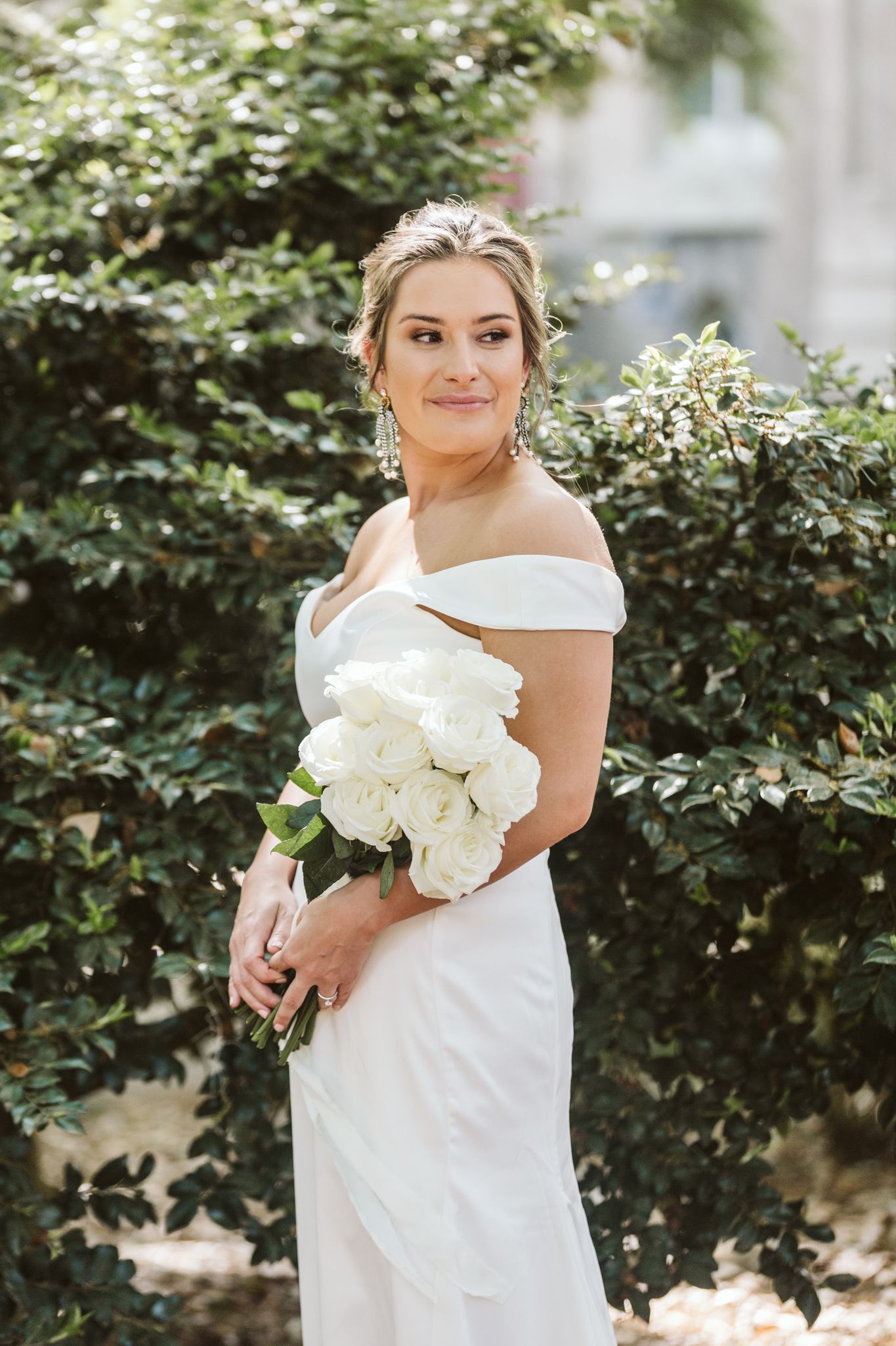 Elegant bride in off-shoulder white gown holds classic white bouquet in garden setting.