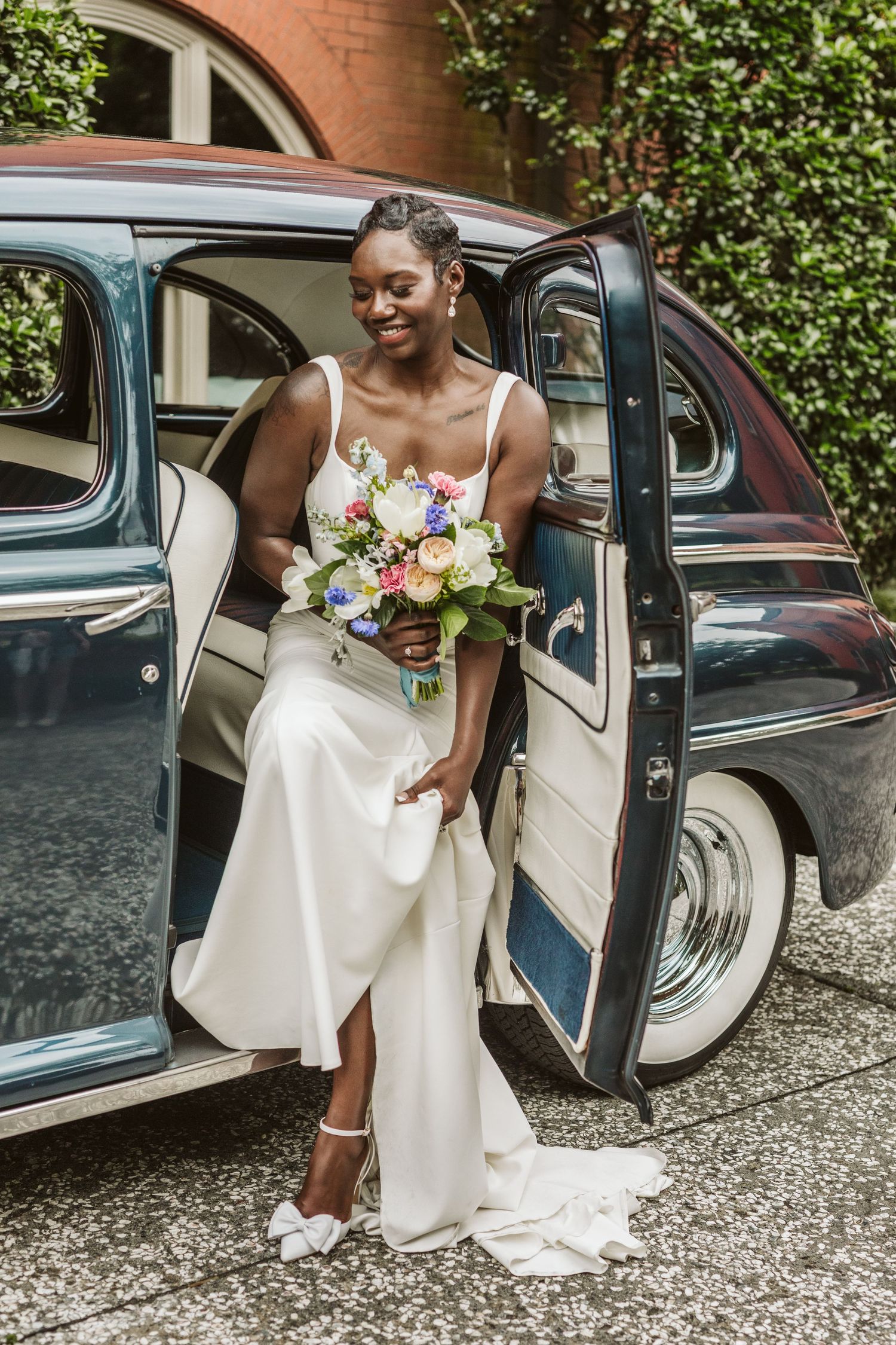 A bride in a white dress elegantly exits a vintage car on her wedding day.