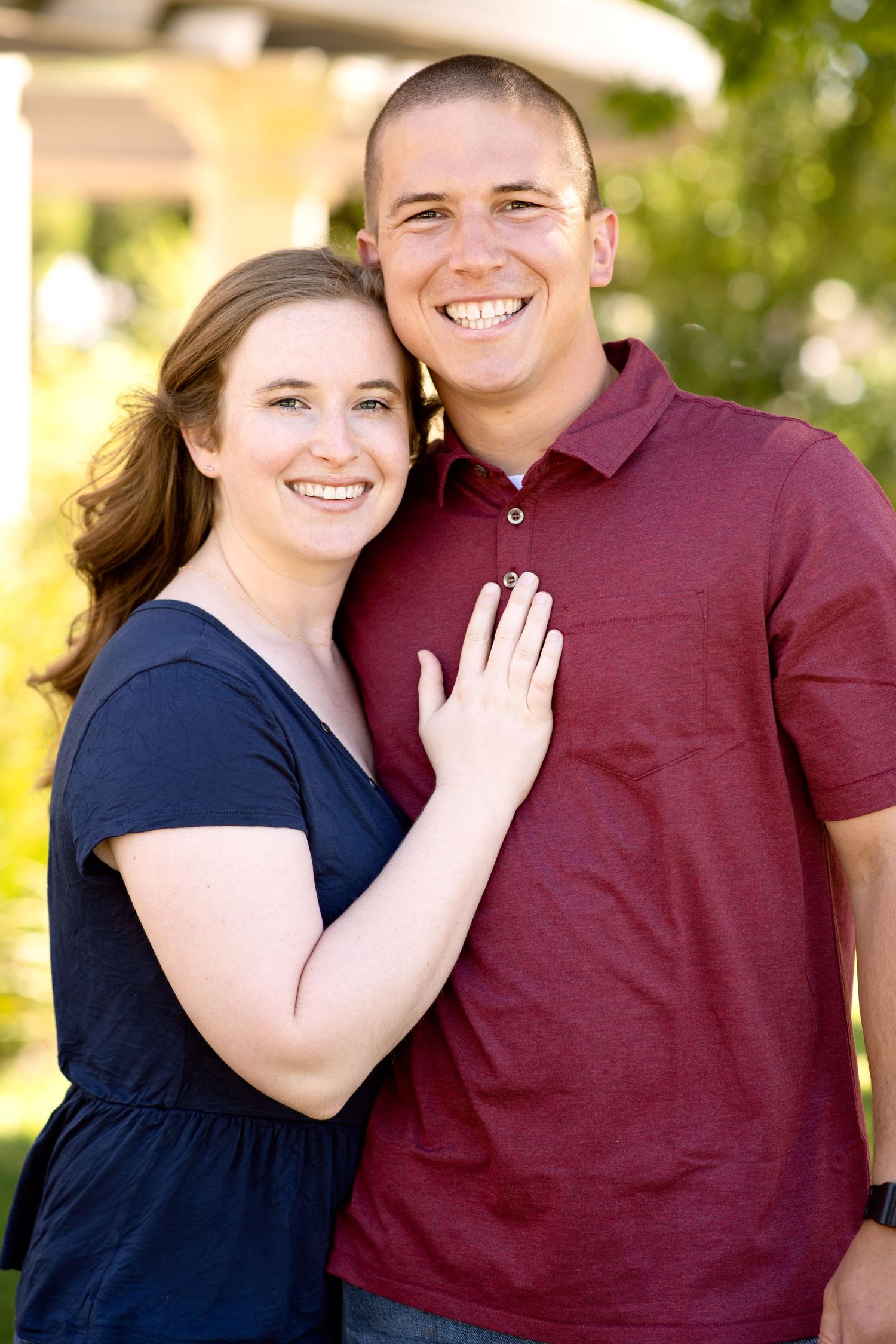 Happy couple in navy dress and burgundy polo shirt embracing outdoors in sunlit setting.