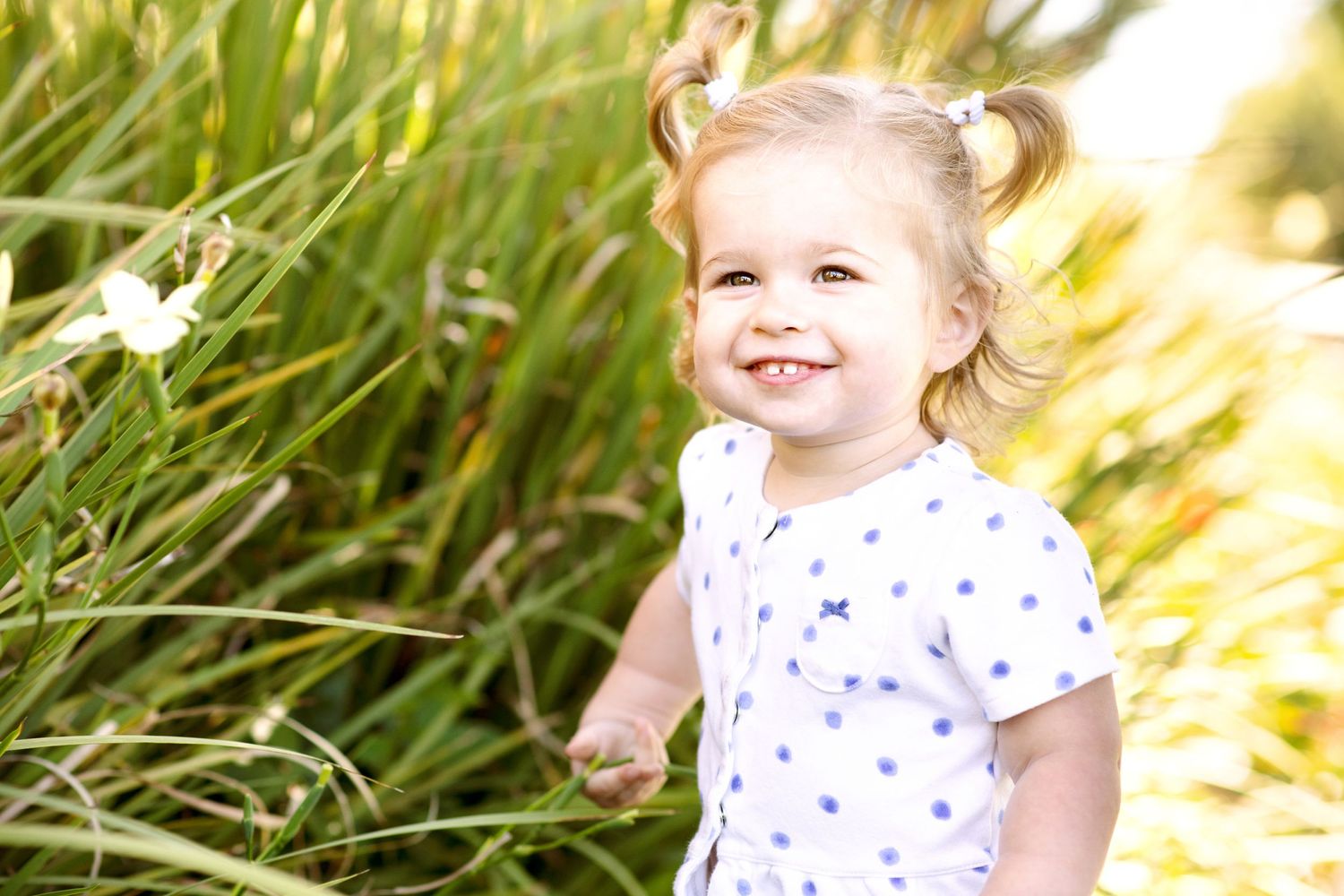 A cheerful toddler in a white and blue polka dot dress stands against a sun-lit backdrop of grass.