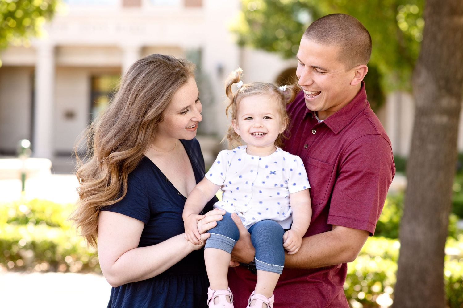 A couple holds their smiling toddler while standing outside on a sunny day.