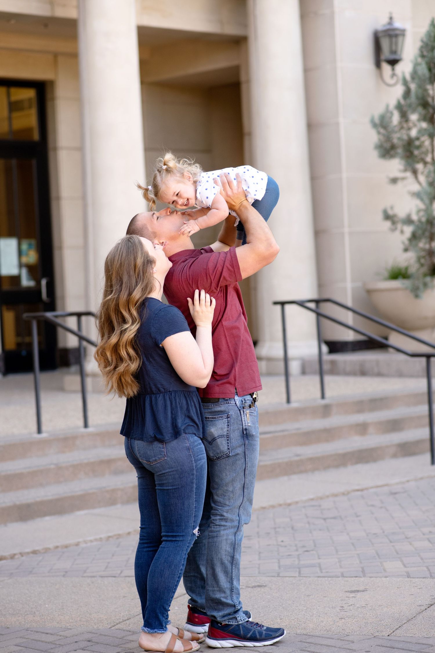 A couple lifts and kisses their toddler while standing on stone steps outside a building.