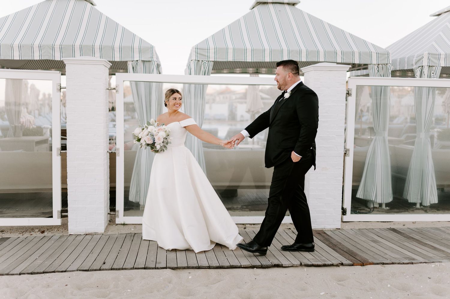 Wedding couple shares romantic moments on a wooden boardwalk with white tent structures in the background.