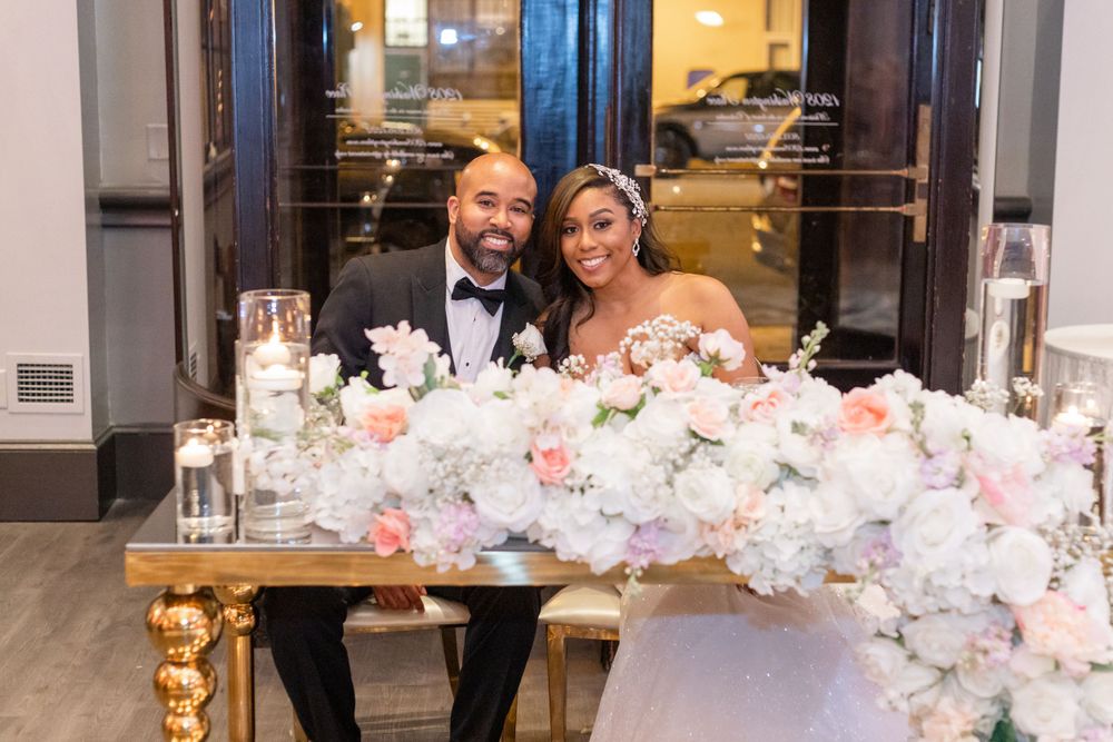 Newlyweds pose at an elegant reception table adorned with white and pink floral arrangements and candles.