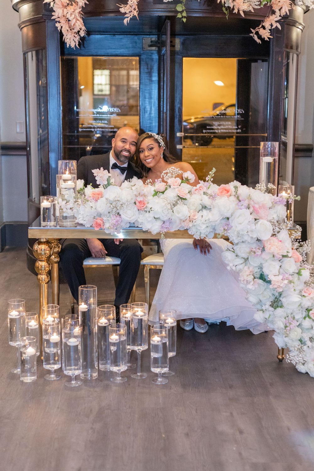 Wedding couple seated at a decorated table with cascading flowers and glowing candles.