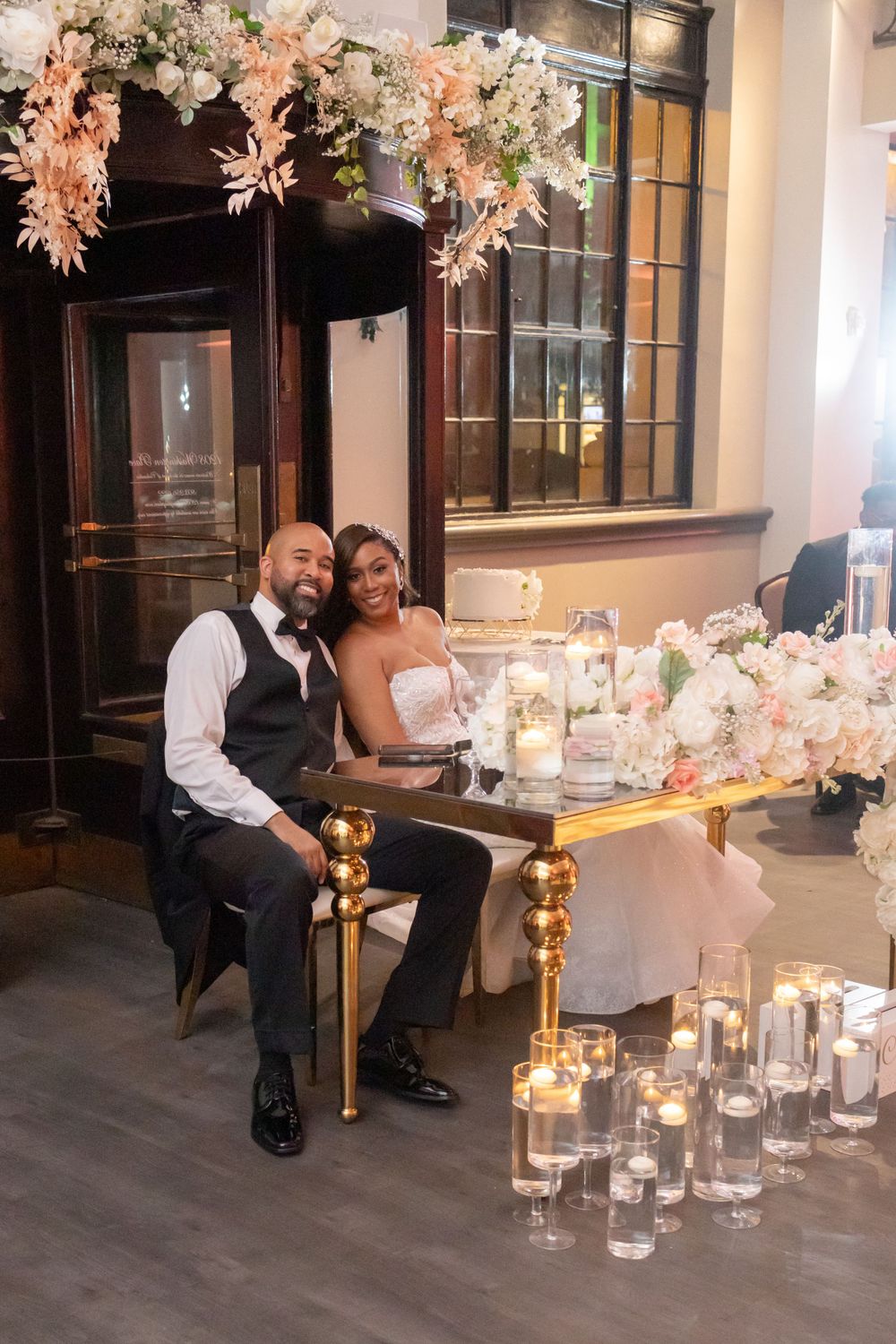 Elegant wedding couple poses at reception table with floral arch and candlelight.