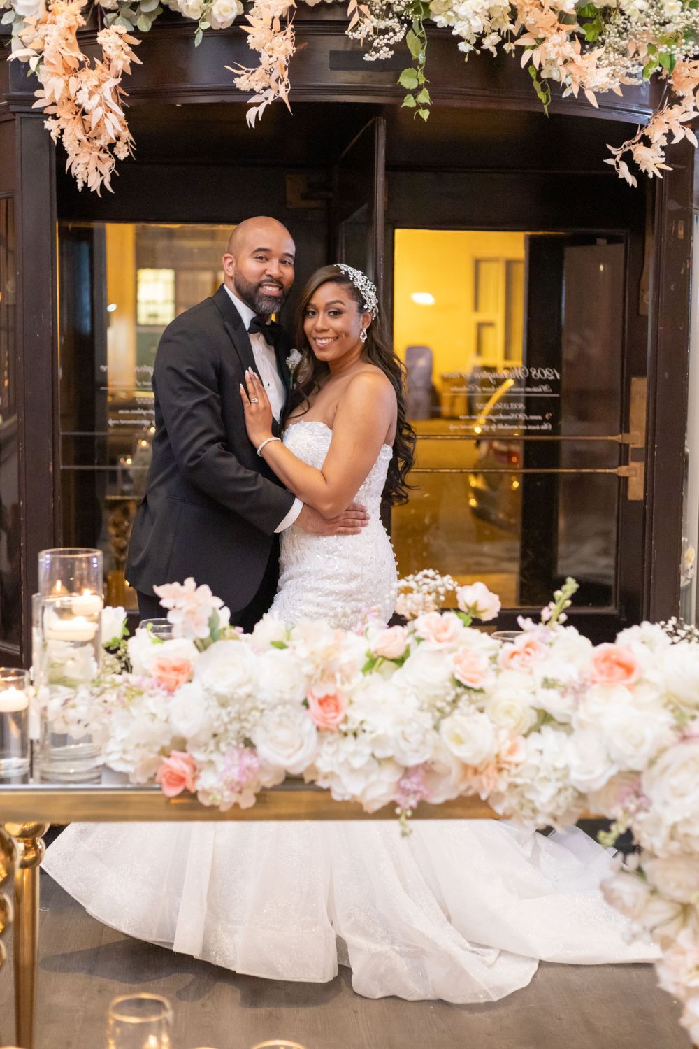 Wedding couple embraces under floral arch with romantic candlelight setting.