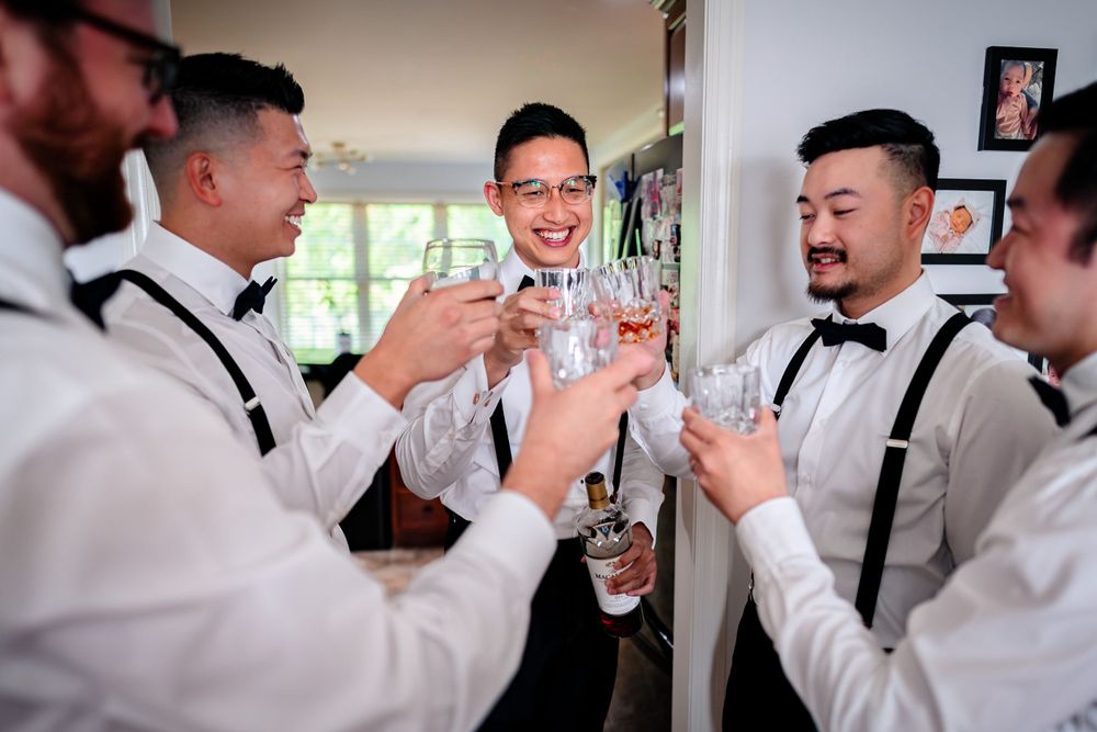 Groomsmen in black suits and bowties share celebratory drinks and laughter during a wedding reception.
