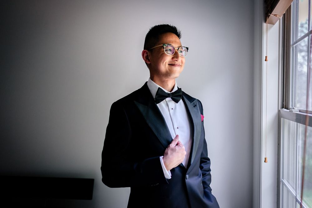 Man in formal black tuxedo and bowtie poses thoughtfully by window in dramatic lighting.