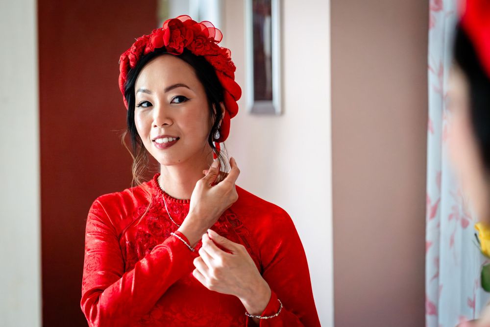Woman in vibrant red traditional dress and floral headpiece smiles during wedding preparations.