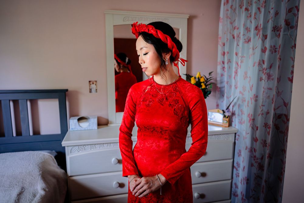 Woman in elegant red lace dress poses in room with floral curtains during wedding preparations.