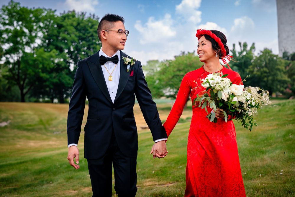 Couple in red dress and black tuxedo walk together on rolling green hillside.