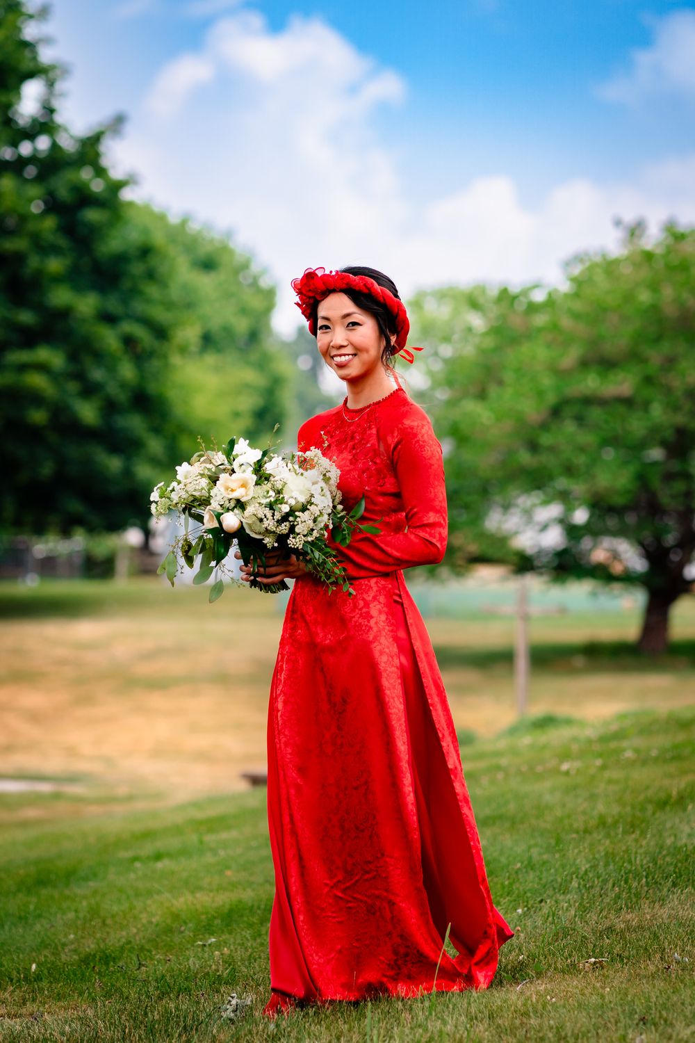 Person in flowing red dress holds white floral bouquet in sunny outdoor setting.