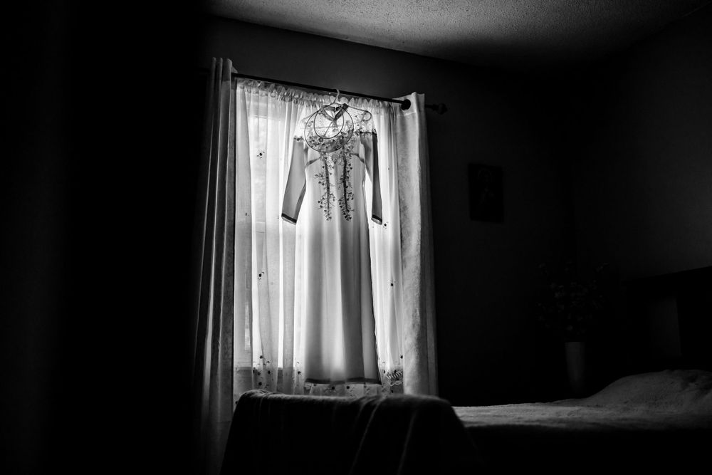 A wedding dress hangs in silhouette against a bright window creating a dramatic lighting effect.