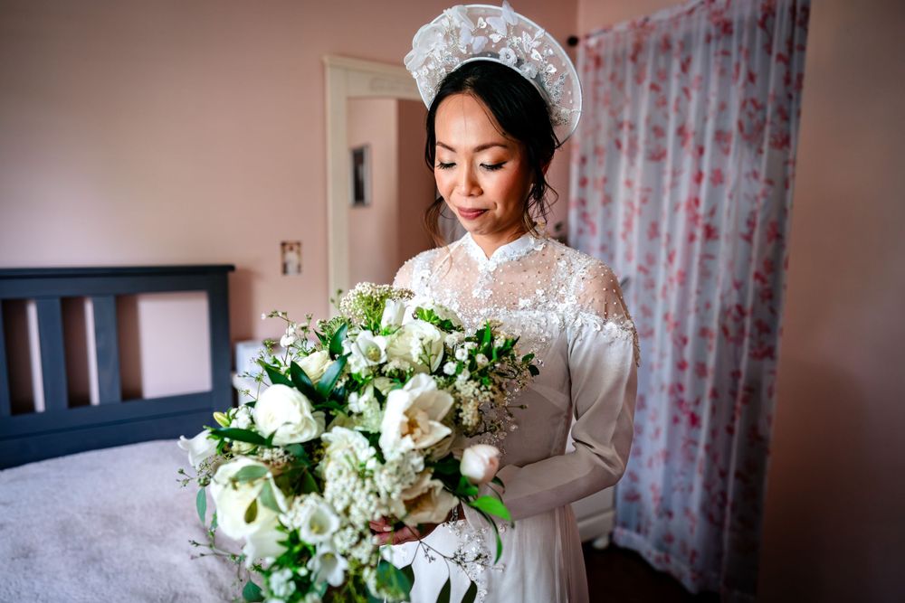 A bride in lace holds a white floral bouquet while getting ready in a pink-tinted room.