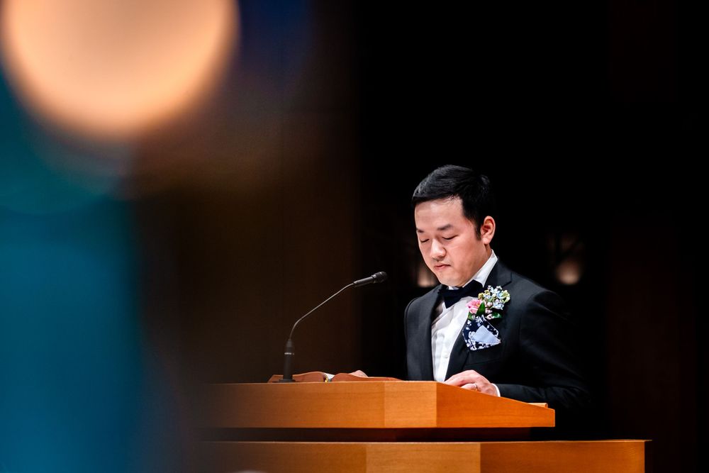 A person speaks at a wooden podium during a dimly lit ceremony.