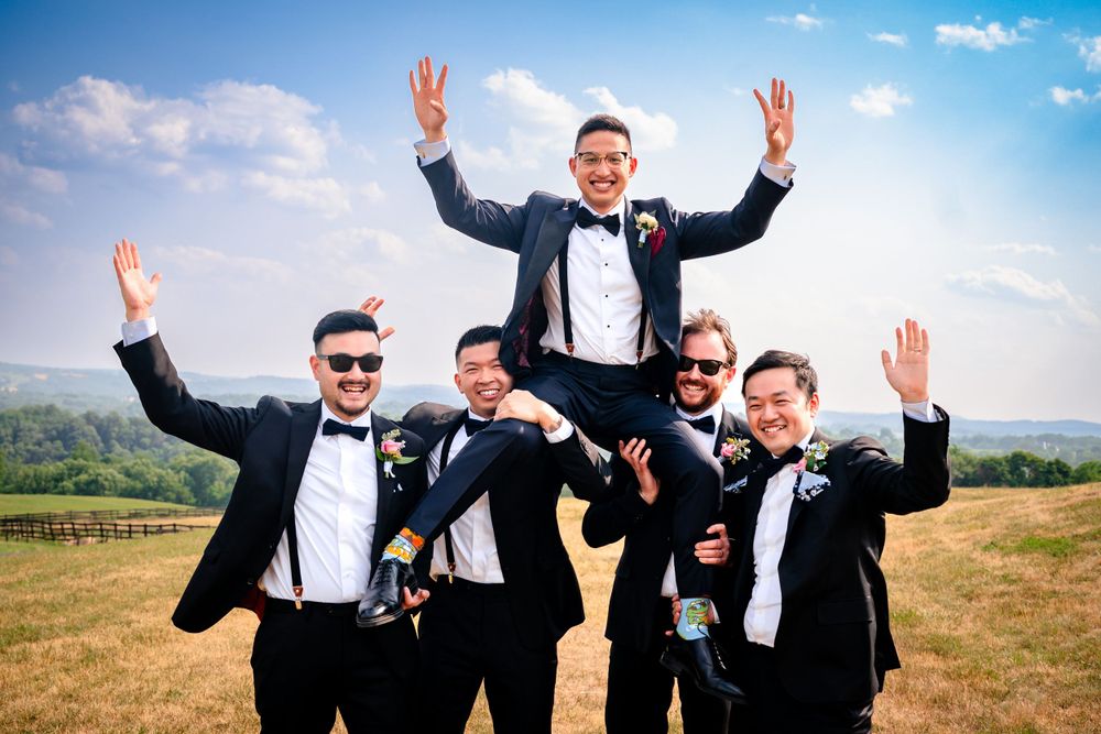 Groomsmen jump and celebrate with enthusiasm on scenic hilltop during wedding festivities.