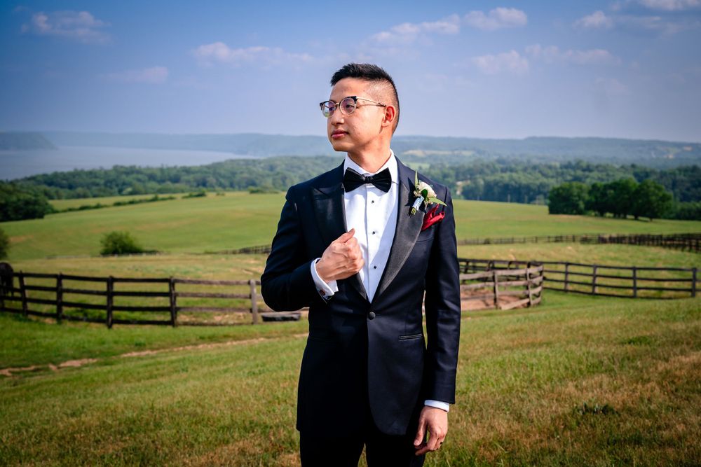 Groomsmen pose playfully in black tuxedos against rustic farm landscape.