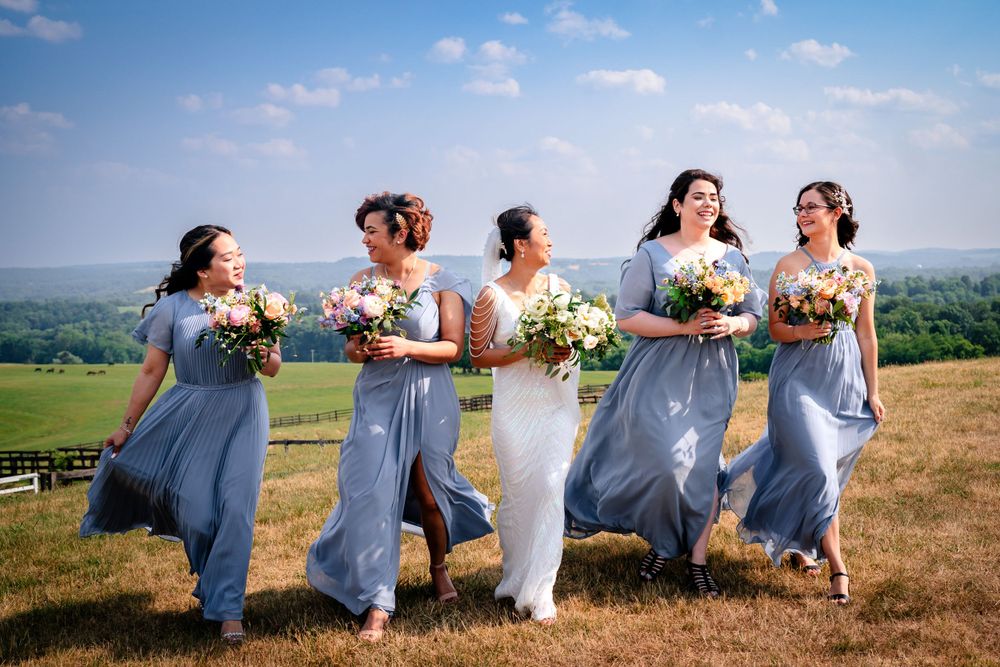 Wedding party in blue dresses laughing and walking together across a scenic hilltop meadow.