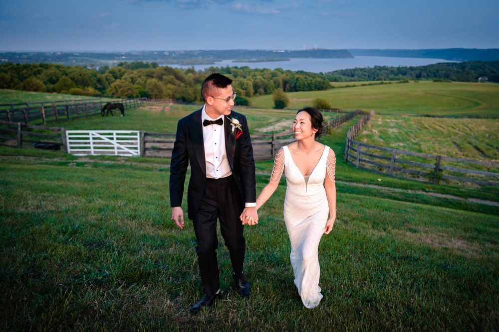 A couple walks hand in hand through a scenic hillside field with white fences in the background.
