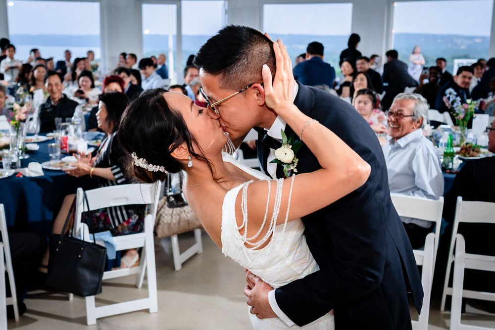 Wedding reception guests celebrate in a bright indoor venue with large windows overlooking water.