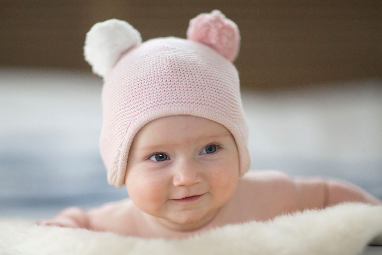 Baby wearing a pink knit hat with pom pom ears lies on soft white blanket.