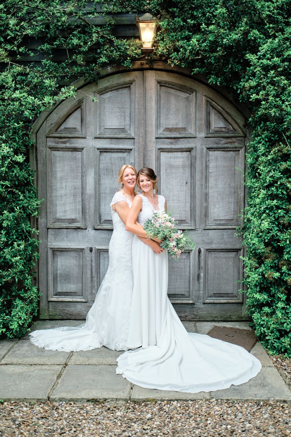 Two people in flowing white dresses pose against an arched wooden door framed by ivy.