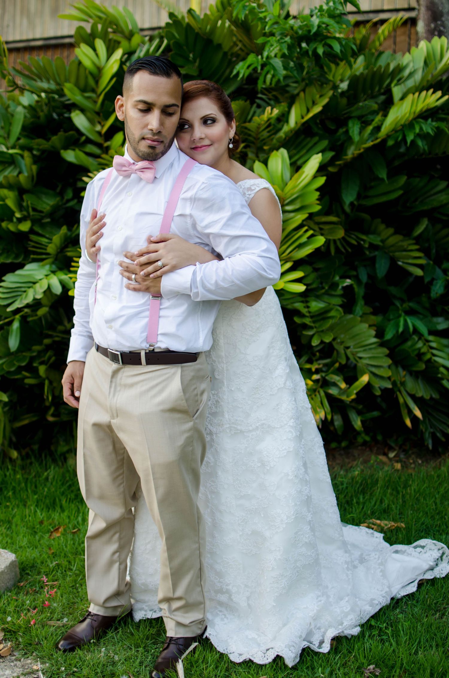 Couple in casual wedding attire embraces in front of lush tropical garden foliage.