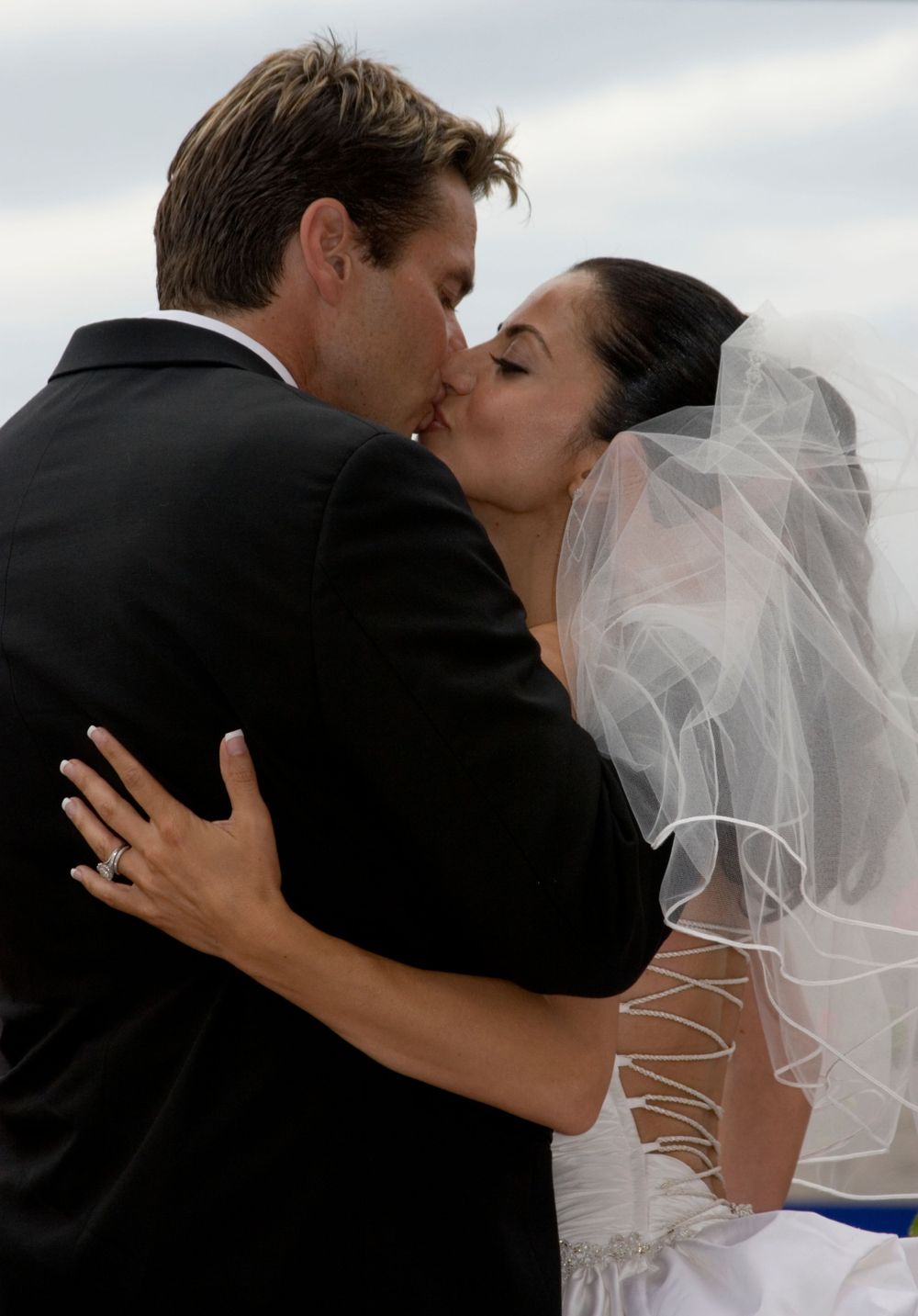 Bride and groom share a passionate kiss outdoors with dramatic cloudy sky in background.