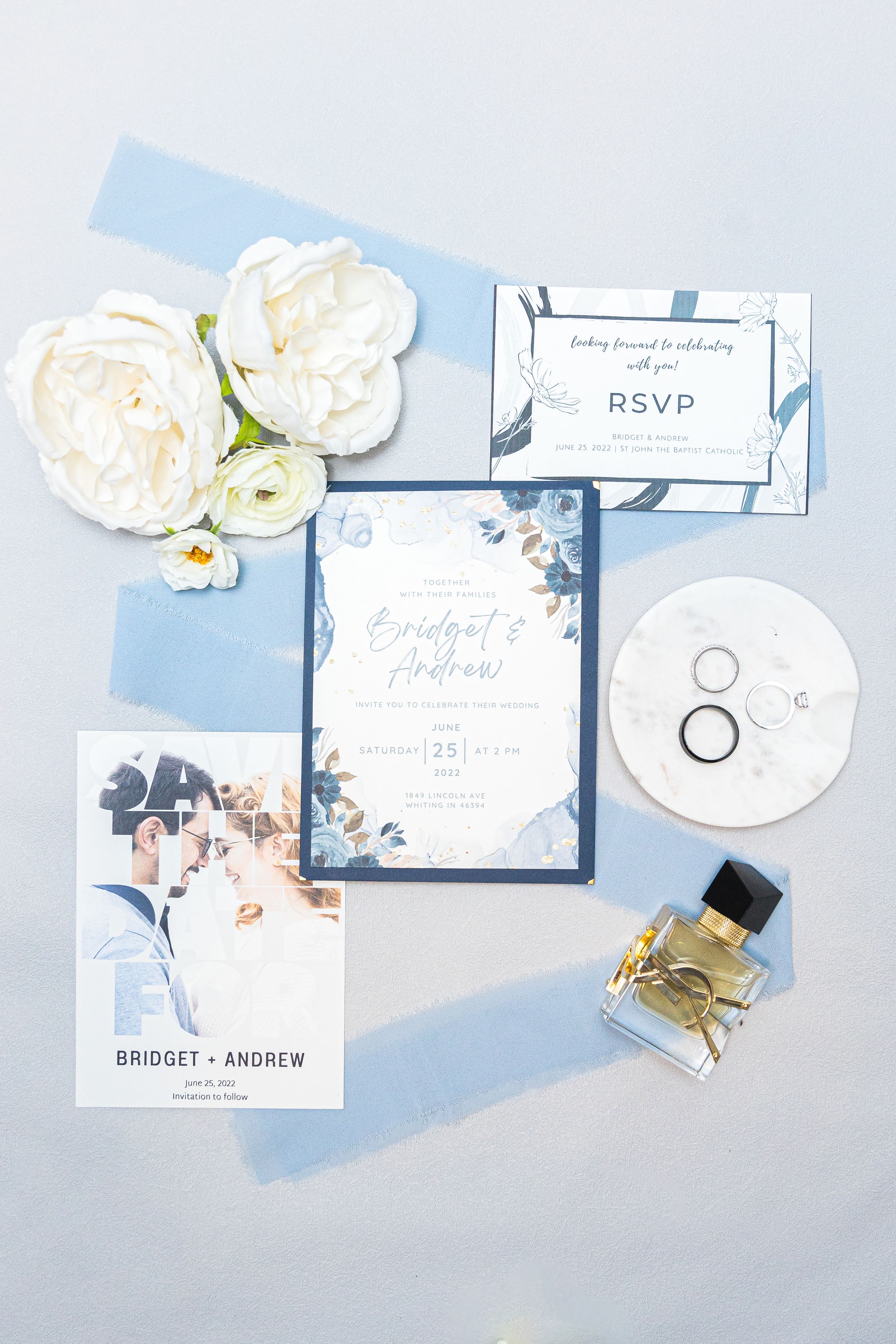 Elegant wedding invitation suite in light blue and white with floral details, perfume bottle and wedding rings on gray surface.