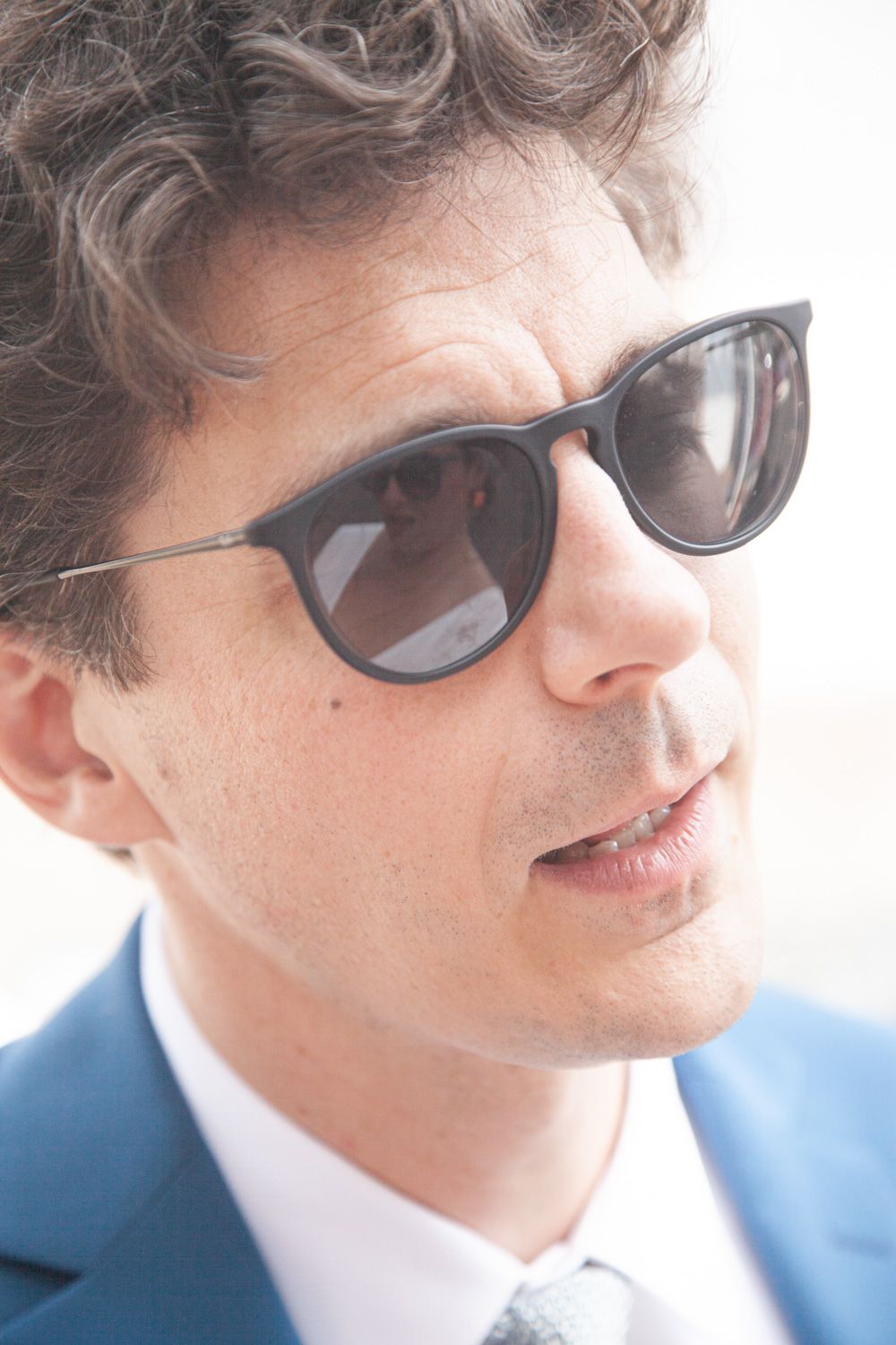 Close-up profile of person wearing sunglasses and blue suit jacket in natural lighting.