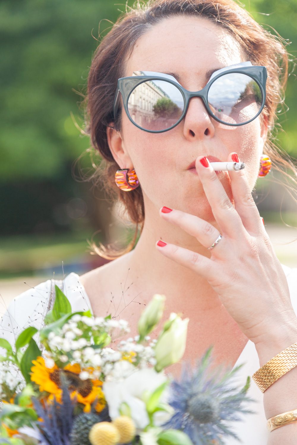 Bride wearing sunglasses and holding a wildflower bouquet while smoking a cigarette.