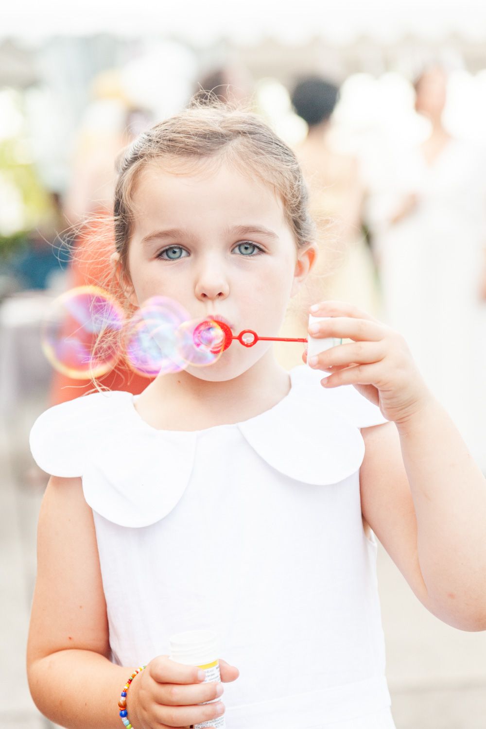 Young child in white dress blowing bubbles outdoors on a sunny day.