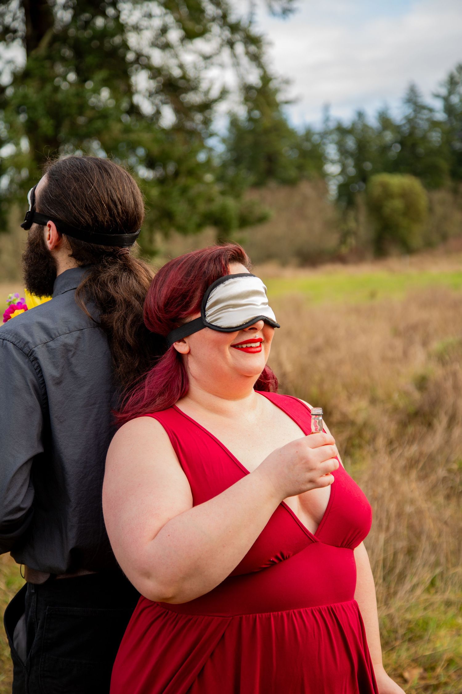 Two people in formal attire share a playful blindfolded moment in a rural field.