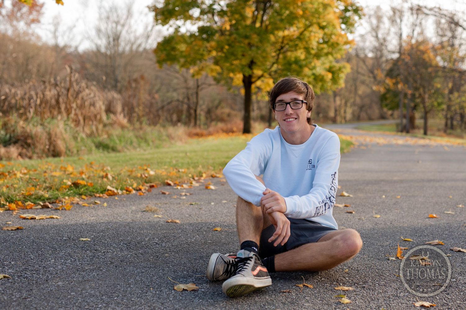 Person in casual attire sits cross-legged on pathway surrounded by fall foliage.